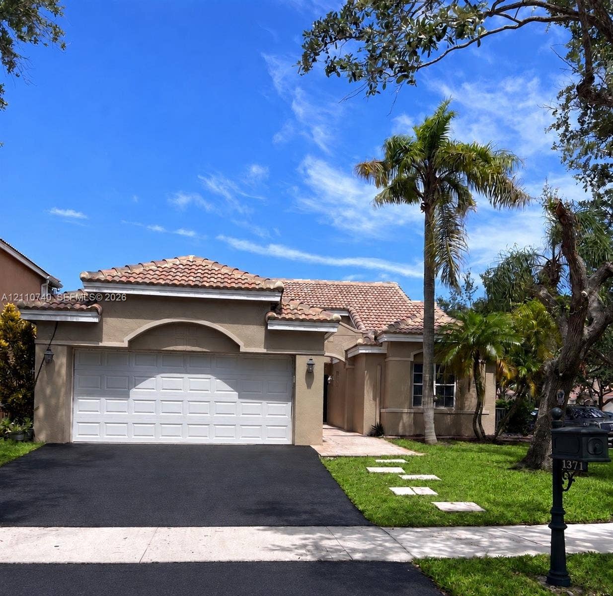 Stucco exterior featuring a tiled hip roof, arched entryway, attached two-car garage, and a landscaped front lawn with mature palm trees