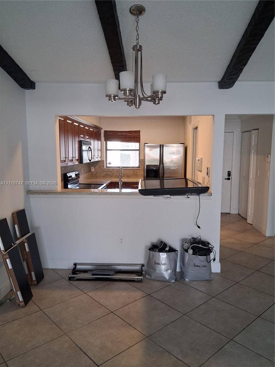 Open-concept interior featuring exposed dark wood-finish ceiling beams, a brushed nickel five-light chandelier, ceramic tile flooring, and a pass-through opening with a stone-finish countertop