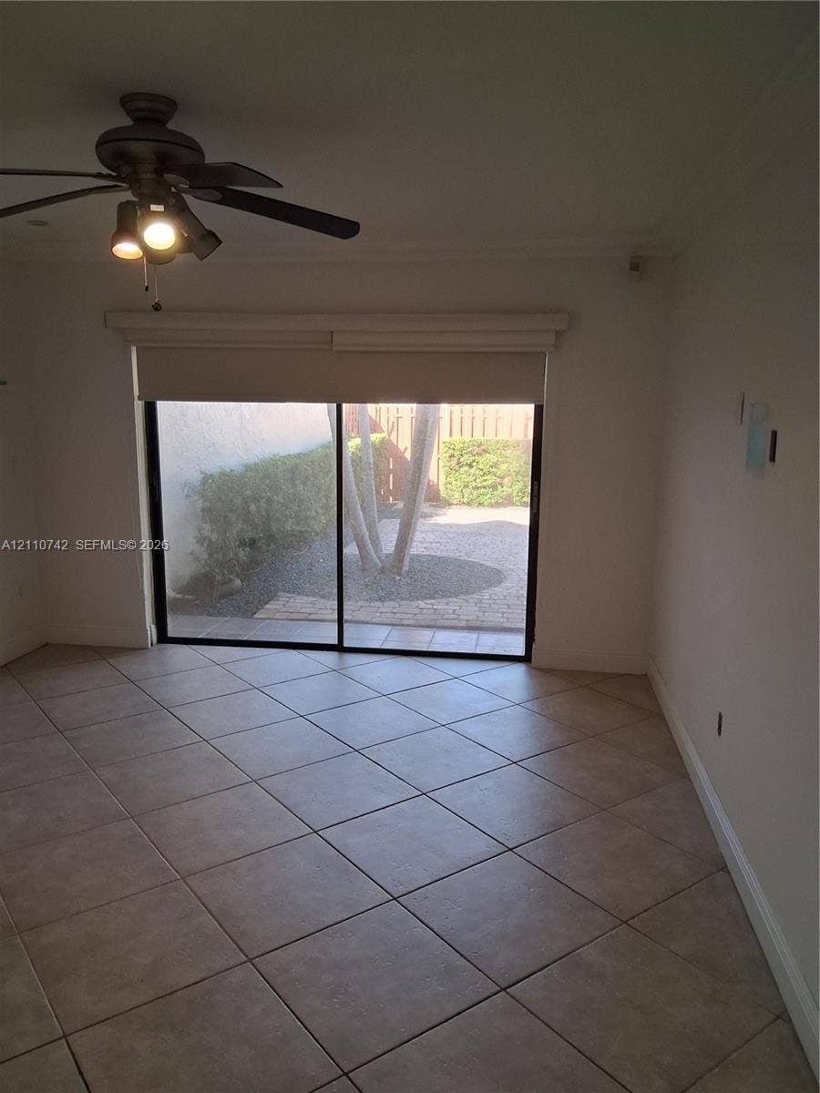 Neutral tile flooring, ceiling fan with integrated lighting, and sliding glass door opening to a paved patio area with mature landscaping