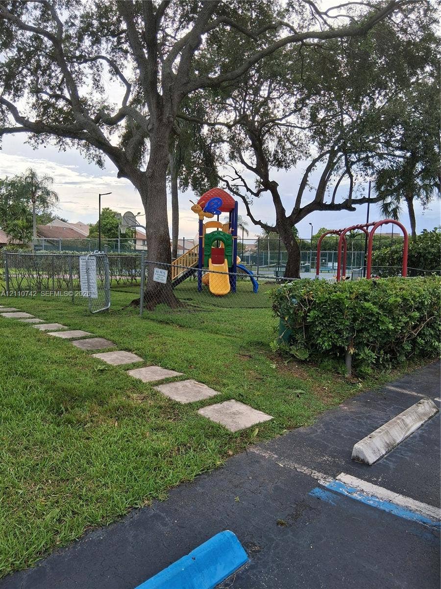 Community recreation area featuring a fenced playground, paved walkways, mature shade trees, and perimeter landscaping
