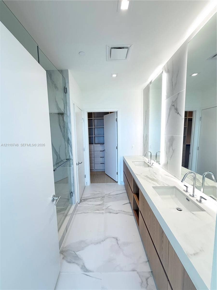 Dual-sink vanity with stone-finish countertop, full-height tile backsplash, glass-enclosed shower, and large-format porcelain tile flooring