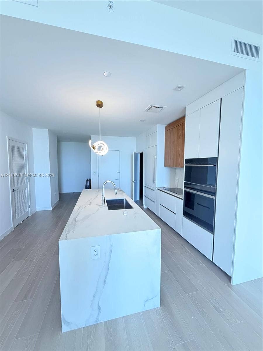 Kitchen featuring a large central island with a waterfall-edge countertop, stone-finish surfacing, integrated sink, and gooseneck faucet, paired with white flat-panel cabinetry, wood-finish accents, and a built-in double oven