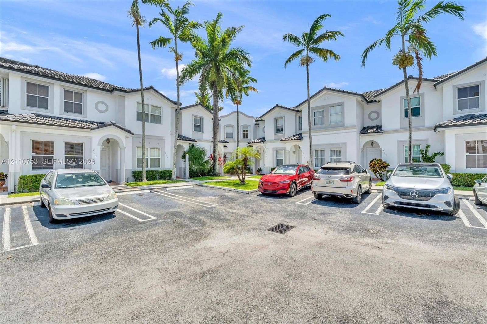 Multi-unit complex featuring light-toned stucco facades, dark-tiled rooflines, dedicated surface parking, and mature palm tree landscaping