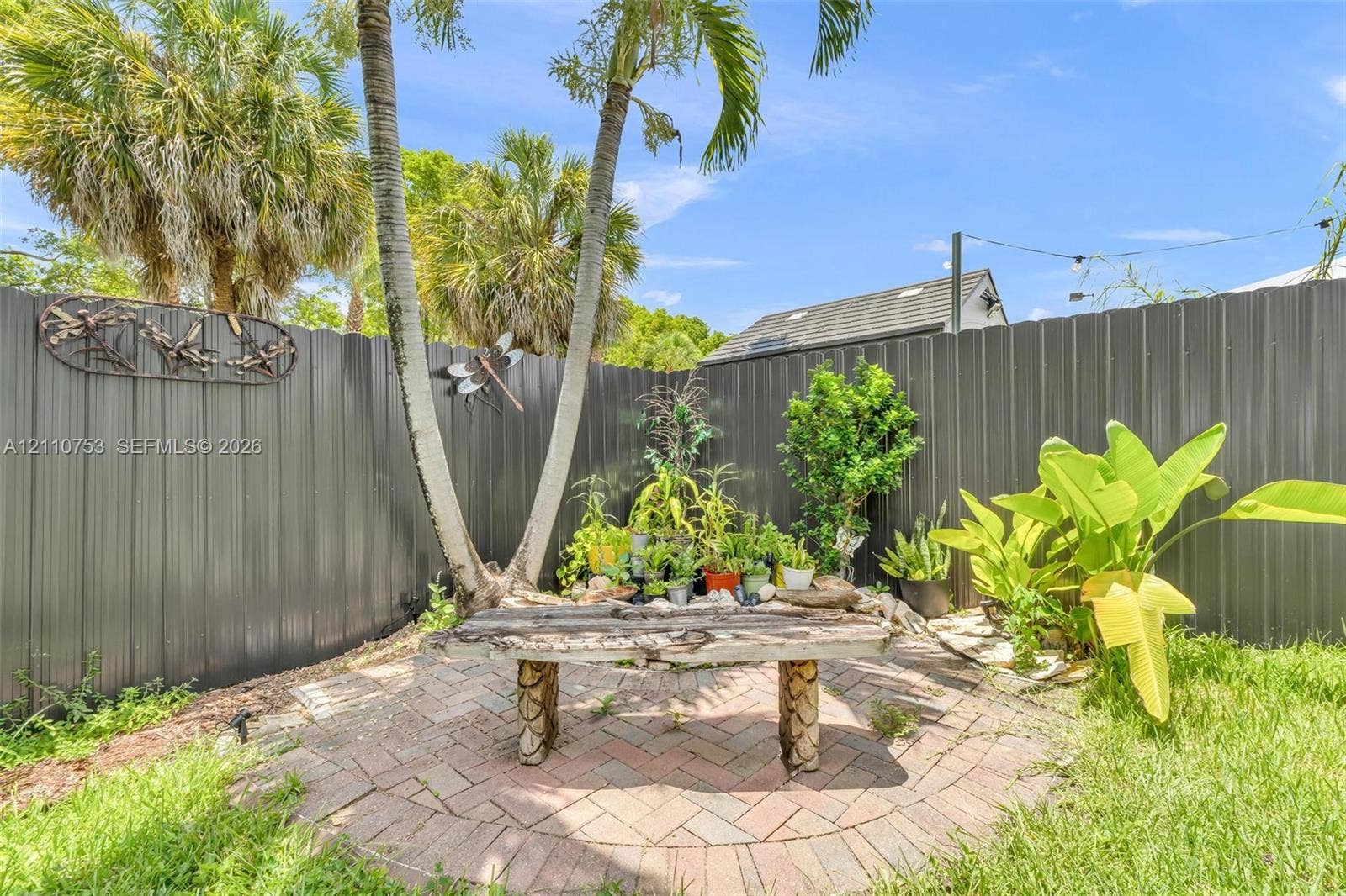 Private backyard featuring a circular brick paver patio, a built-in stone-base bench, tall palms, mature landscaping, and corrugated metal fencing