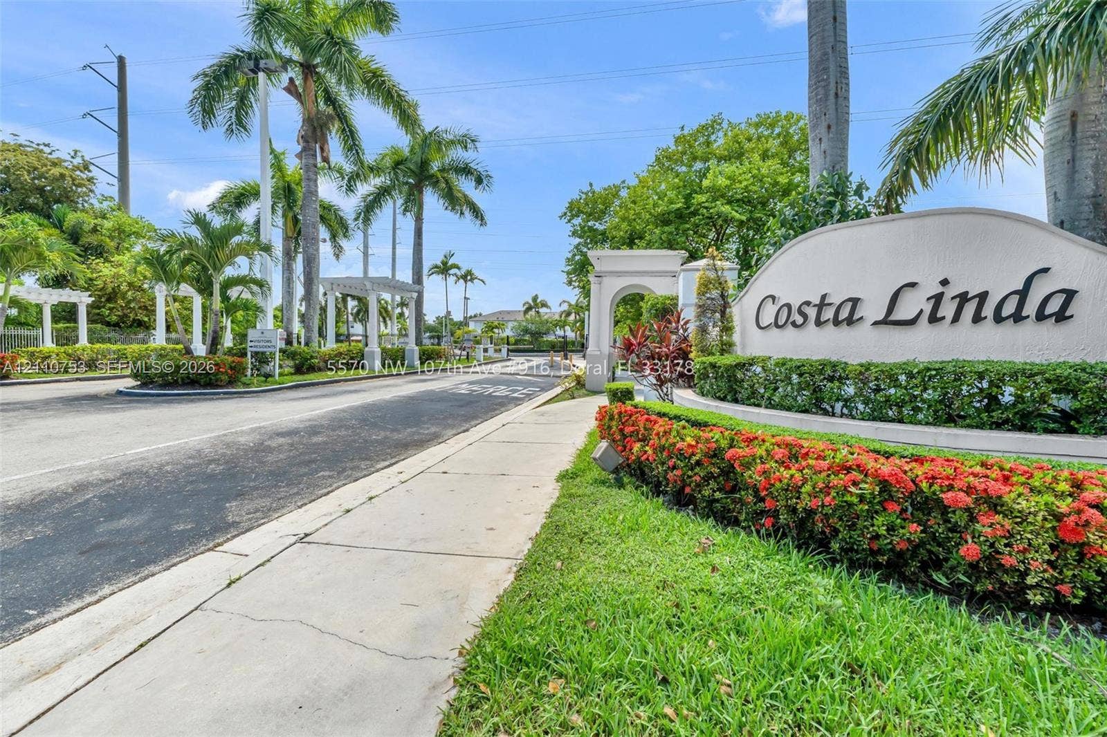 Community entrance featuring arched masonry architecture, paved roadways, mature palm trees, and manicured shrubbery