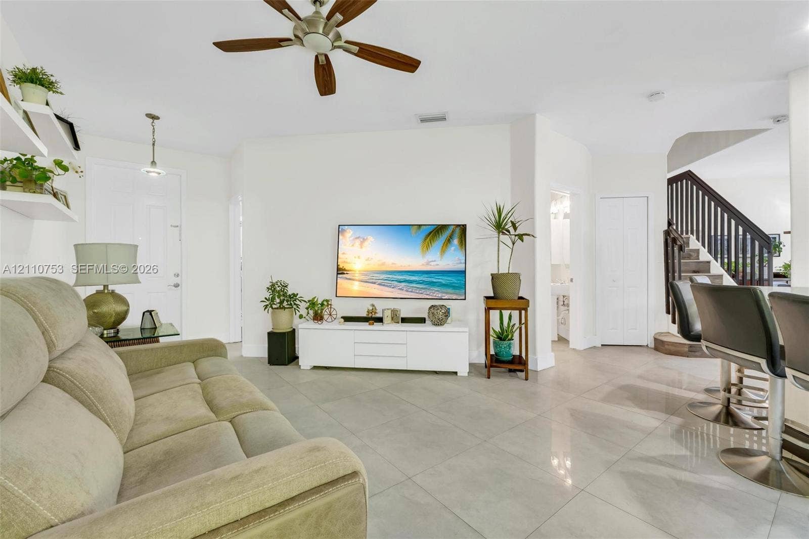 Living area featuring large-format ceramic tile flooring, a multi-blade ceiling fan with integrated lighting, wall-mounted shelving, and a staircase with decorative dark wood railings