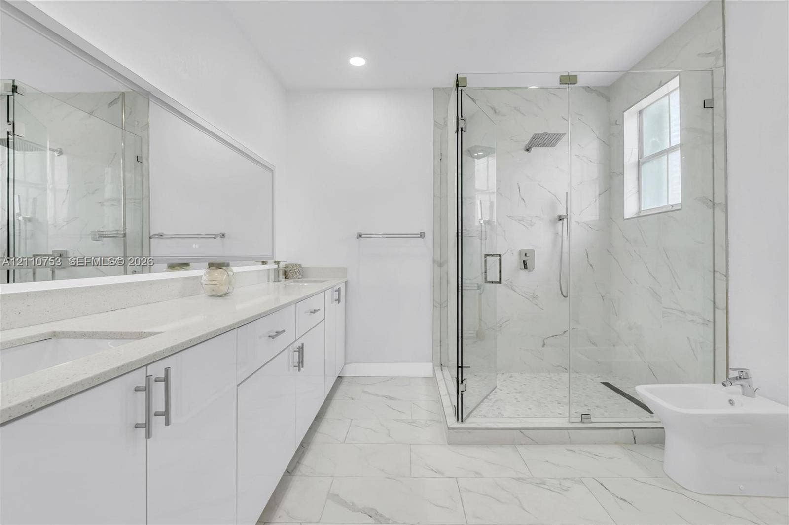 Bathroom featuring a double-sink vanity with light-toned solid surface countertops, floor-to-ceiling tiled glass-enclosed shower, large wall-mounted mirror, and tile flooring