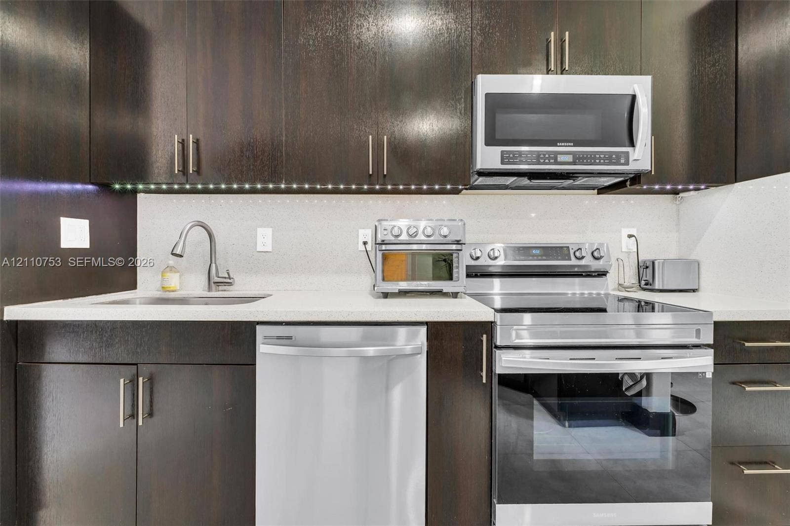 Kitchen featuring dark wood-finish cabinetry, solid surface countertops, stainless steel appliances, under-cabinet lighting, and a gooseneck faucet