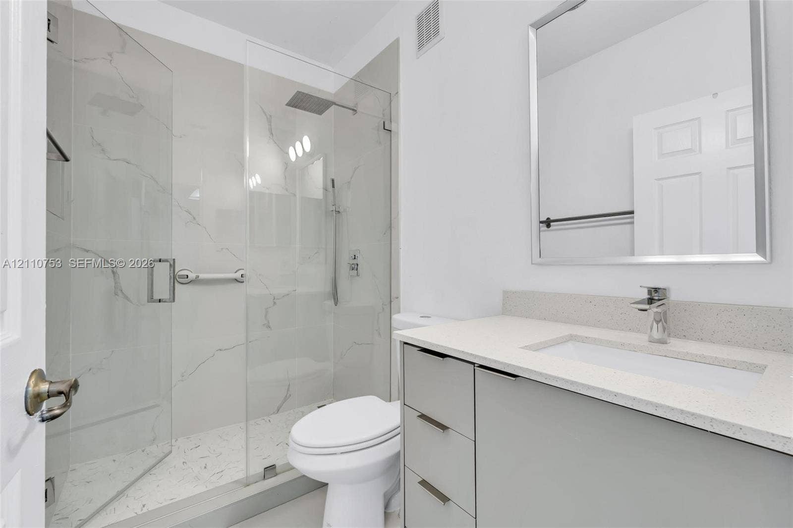 Modern bathroom featuring a light gray vanity with a stone-composite countertop, undermount sink, walk-in shower with floor-to-ceiling large-format tile, frameless glass enclosure, and a silver-framed mirror