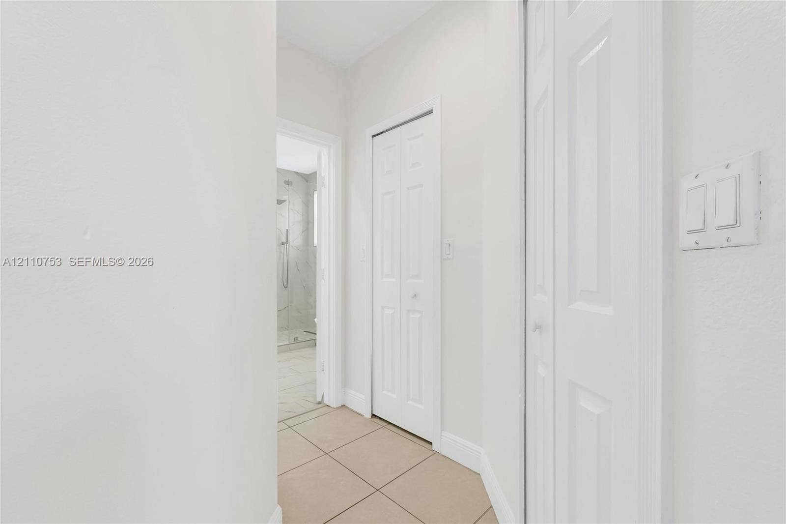 Hallway featuring neutral-toned tile flooring, two bi-fold doors, and a doorway revealing a tiled shower enclosure