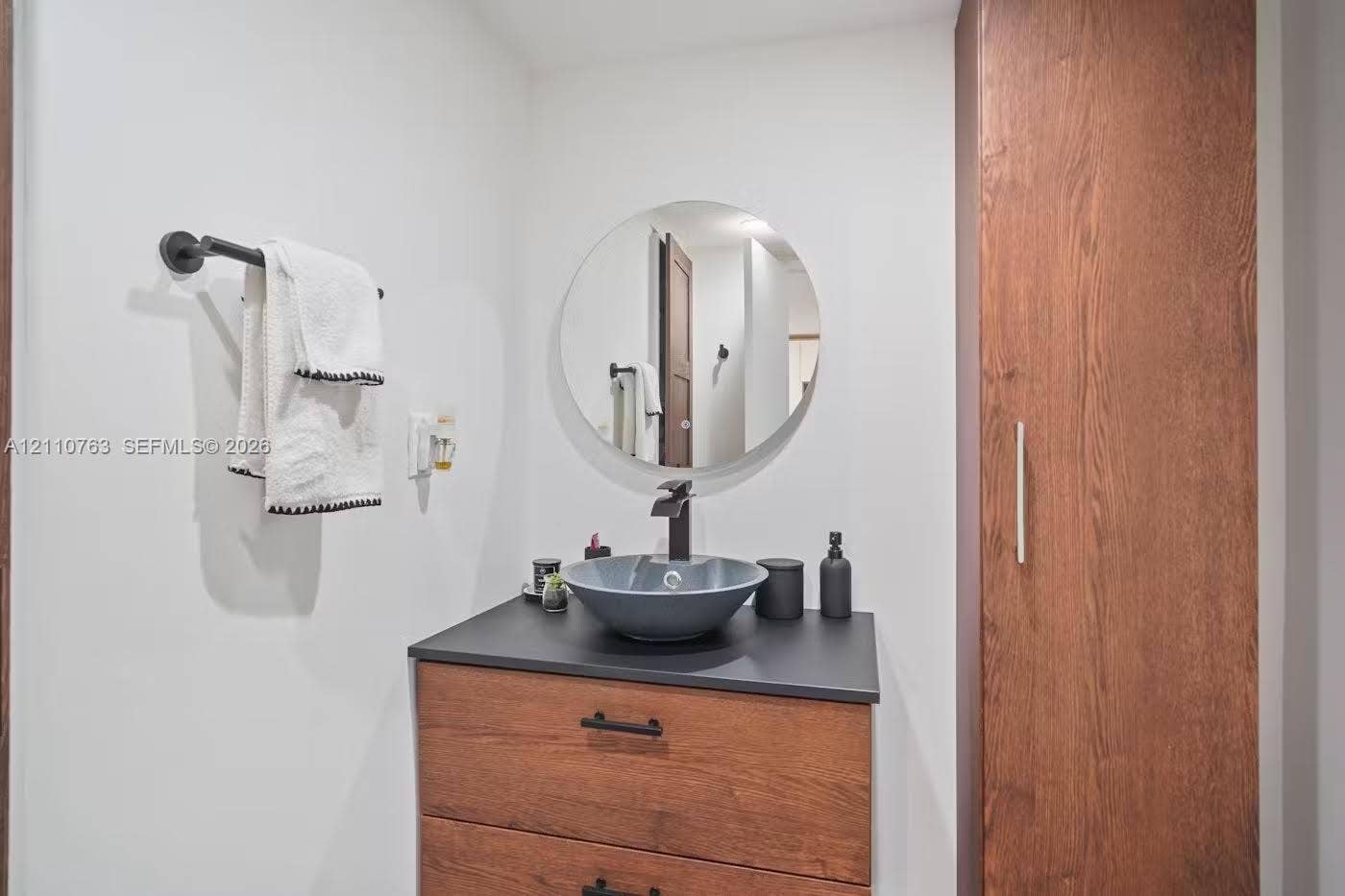 1901 Brickell Avenue, #B1706, Miami, FL 33129 Modern vanity featuring a wood-finish cabinet, dark solid-surface countertop, circular vessel sink, and matte black faucet, complemented by a tall built-in storage cabinet and a circular wall-mounted mirror