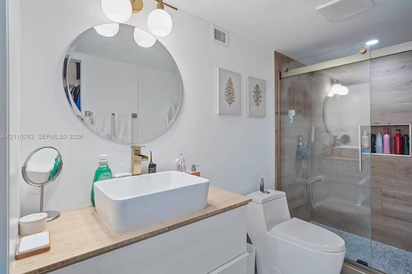 1901 Brickell Avenue, #B1706, Miami, FL 33129 Floating vanity with a light wood-finish countertop, rectangular vessel sink, gold-tone faucet, round wall-mounted mirror, and glass-enclosed shower with wood-finish tile wall and built-in recessed niche