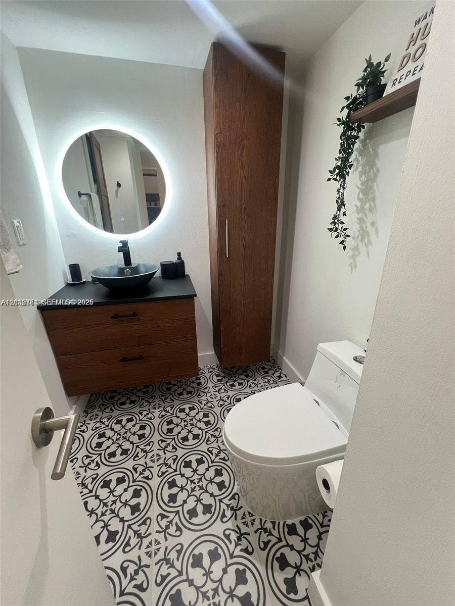 1901 Brickell Avenue, #B1706, Miami, FL 33129 Modern vanity with wood-finish cabinetry, black stone countertop, and circular vessel sink, complemented by a backlit round mirror, floor-to-ceiling wood-finish storage cabinet, and patterned tile flooring