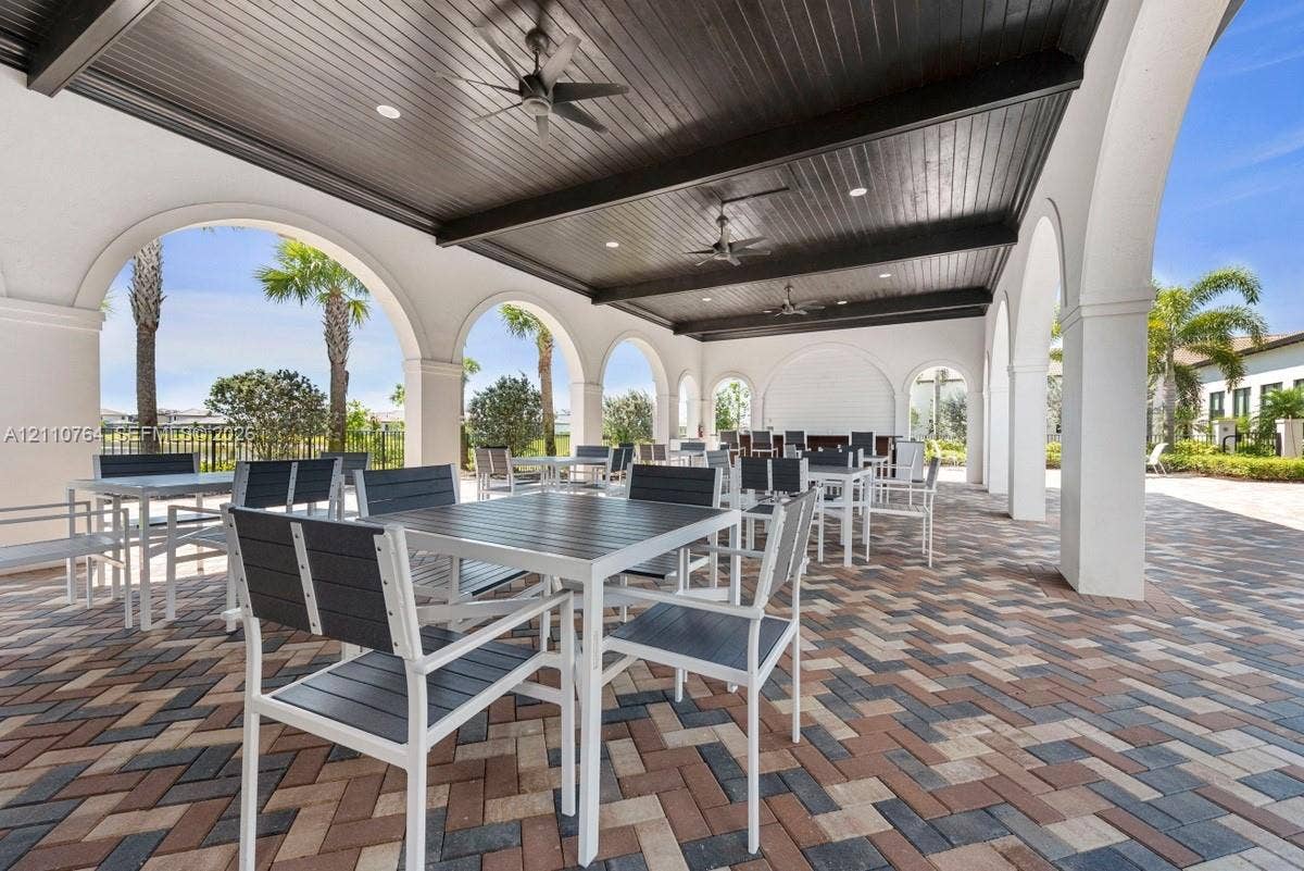 Covered outdoor loggia featuring architectural arched columns, dark wood-plank ceiling, recessed lighting, ceiling fans, and multi-tone herringbone paver flooring