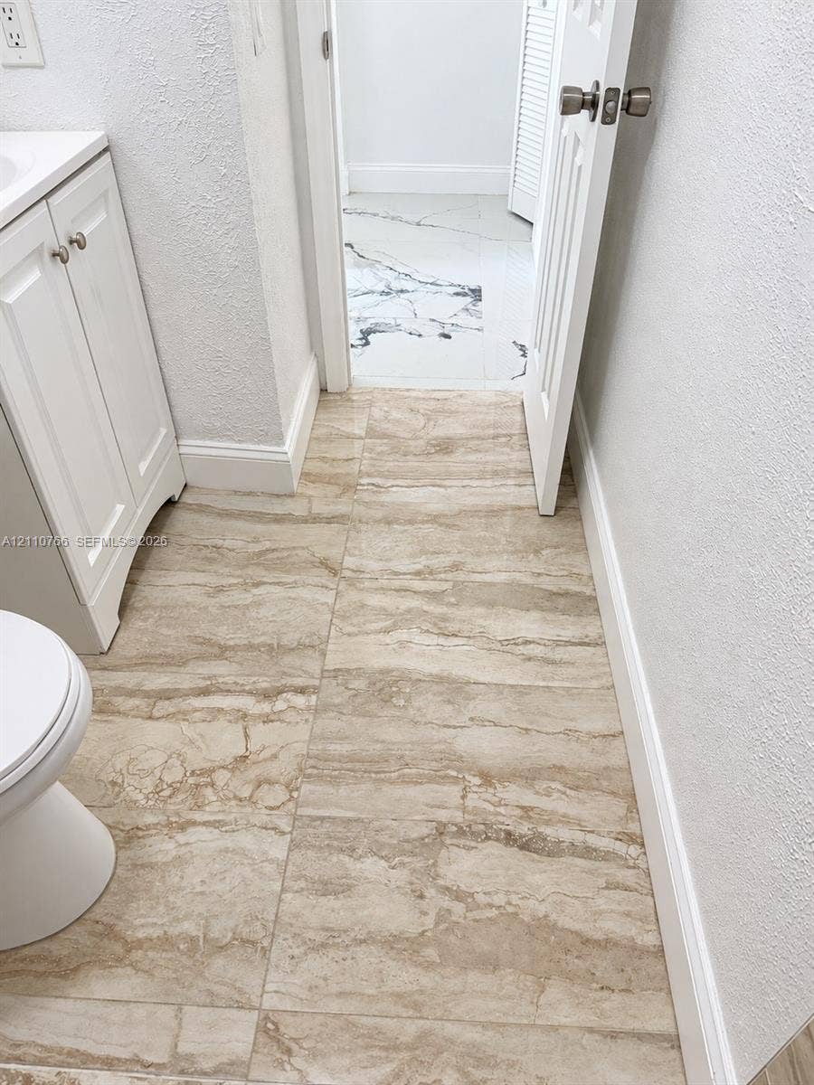 Large-format neutral stone-patterned tile flooring, white shaker-style cabinetry, and contrasting monochromatic veined stone-patterned floor tile visible through the doorway
