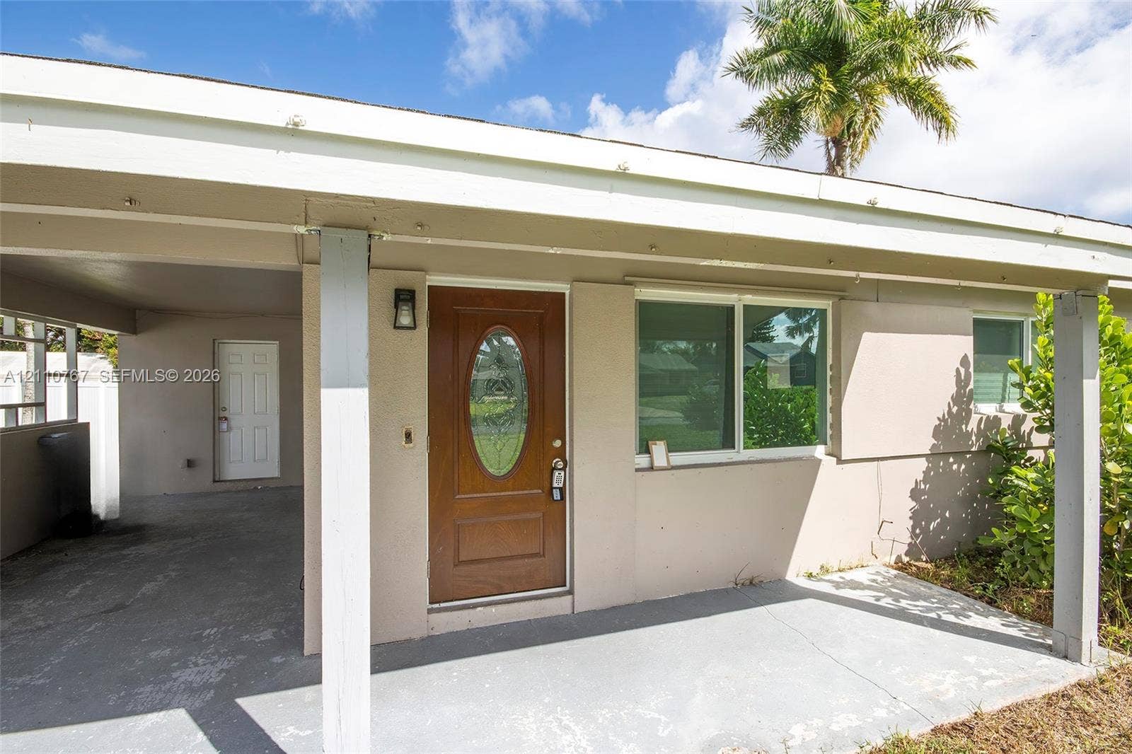 5305 Cleveland Road, Delray Beach, FL 33484 Exterior entryway featuring a covered carport, wood-finished front door with an oval glass inlay, sliding window with horizontal muntins, and a concrete porch