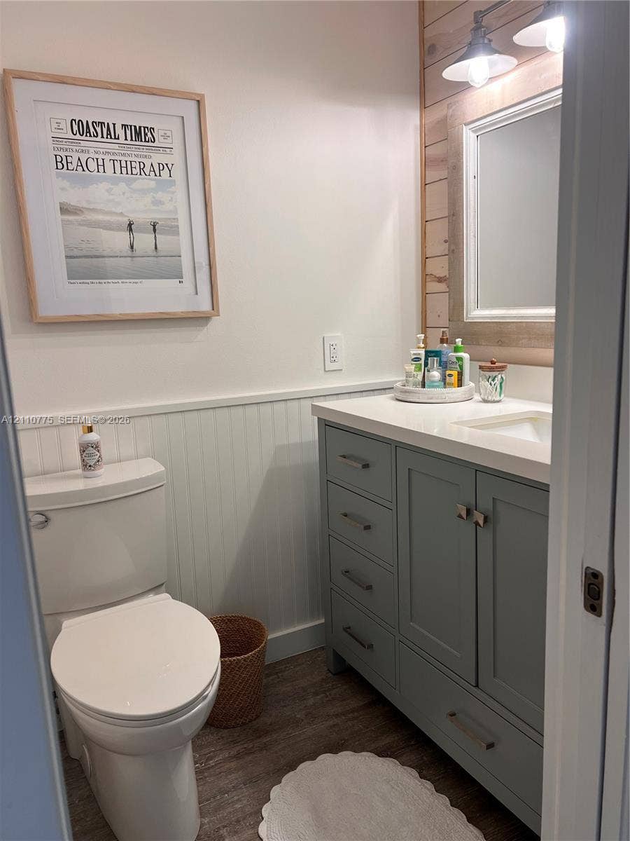 4995 S Highway A1a, #D, Melbourne Beach, FL 32951 Bathroom featuring a vanity with soft-toned cabinetry, light-colored solid surface countertop, white beadboard wainscoting, wood-finish flooring, and horizontal wood-plank accent wall