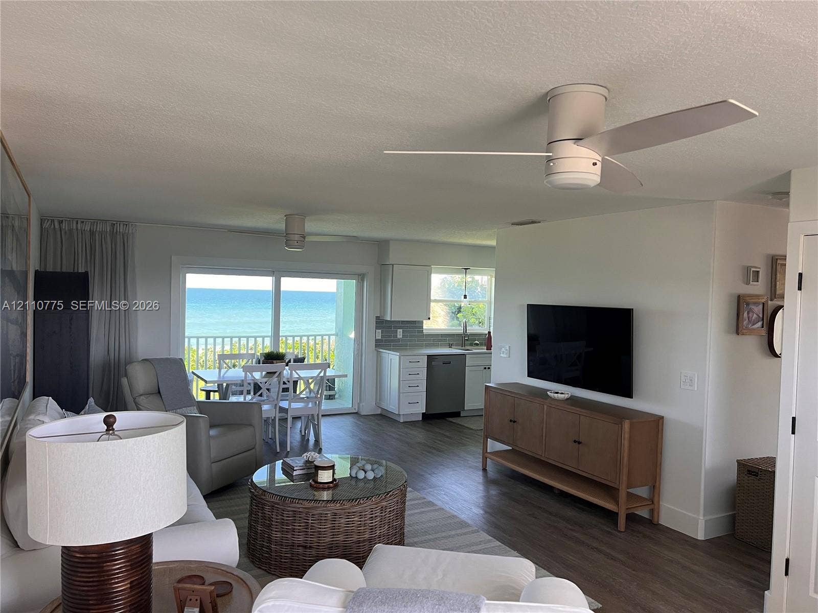 4995 S Highway A1a, #D, Melbourne Beach, FL 32951 Open-concept living space featuring wood-finish flooring, white kitchen cabinetry with tile backsplash, ceiling fans, and large glass sliding doors providing water scenery