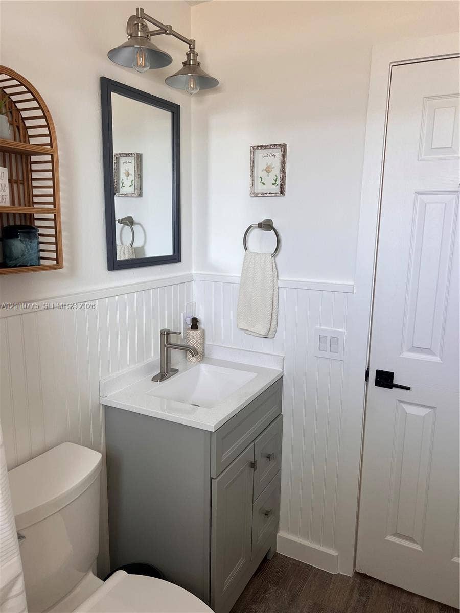 4995 S Highway A1a, #D, Melbourne Beach, FL 32951 Gray vanity with a white countertop and basin, white beadboard wainscoting, dual-light metal vanity fixture, framed wall mirror, and six-panel interior door with matte black hardware