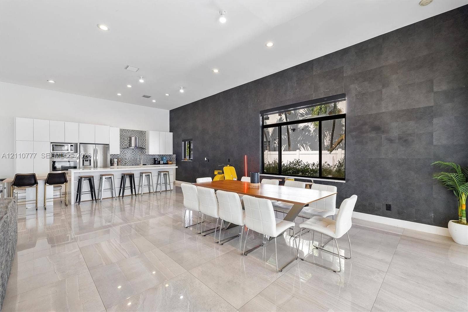 Modern kitchen and dining area featuring high-gloss white cabinetry, expansive stone tile flooring, a dark charcoal textured accent wall, recessed lighting, and built-in stainless steel wall ovens