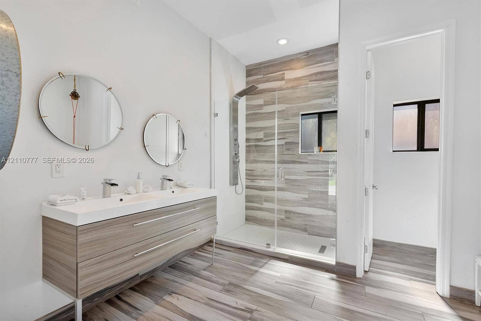 Double vanity with integrated sinks, wall-mounted faucets, wood-finish cabinetry, walk-in shower with horizontal tile surround, and wood-finish flooring
