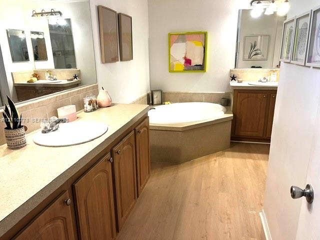 Dual vanities featuring wood-finish cabinetry, solid-surface countertops, integrated sinks, and expansive wall-mounted mirrors, complemented by a built-in soaking tub and wood-finish flooring