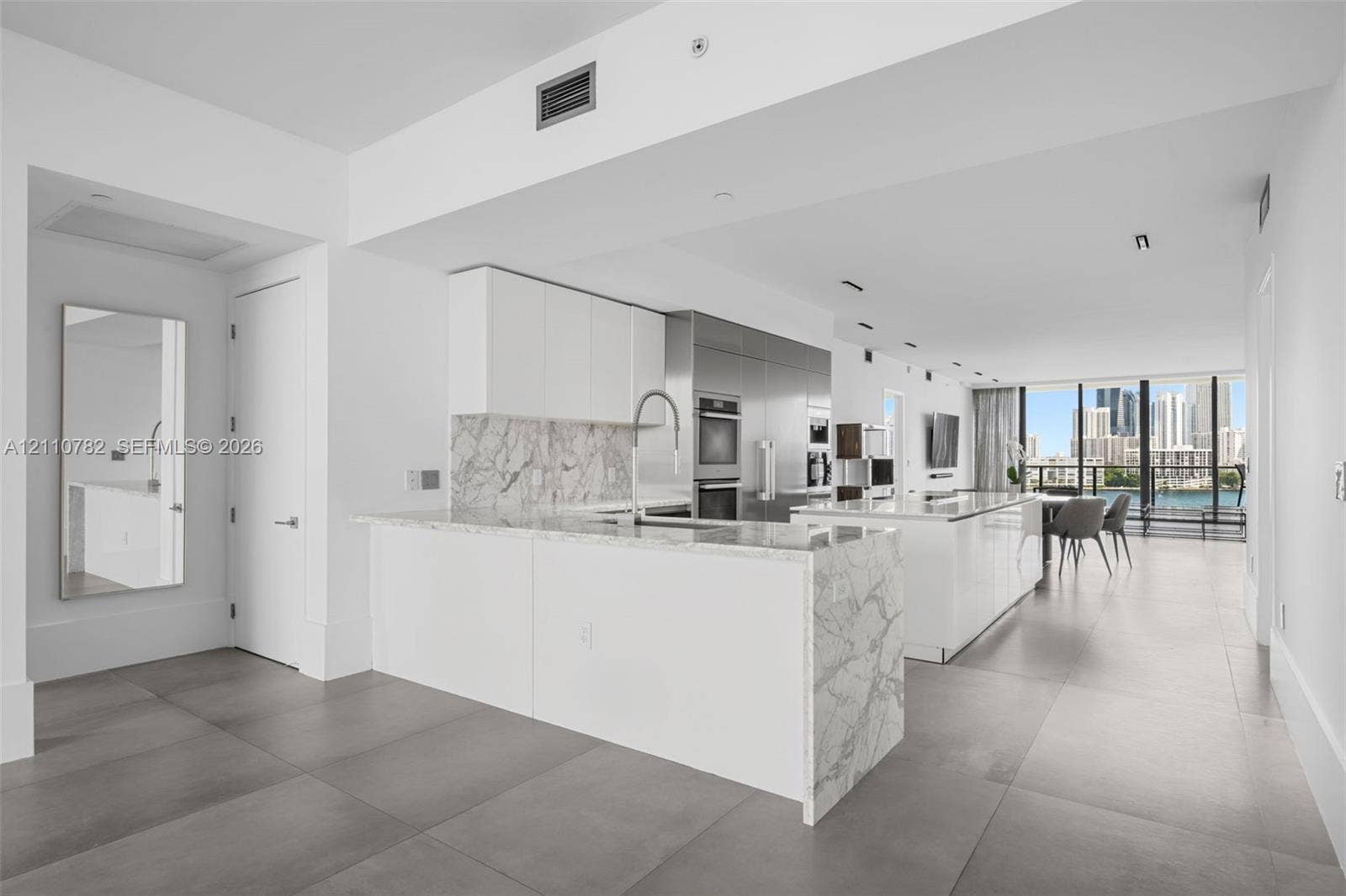 Kitchen featuring a stone-finish island with waterfall edge, white flat-panel cabinetry, stainless steel built-in appliances, and large-format floor tiling