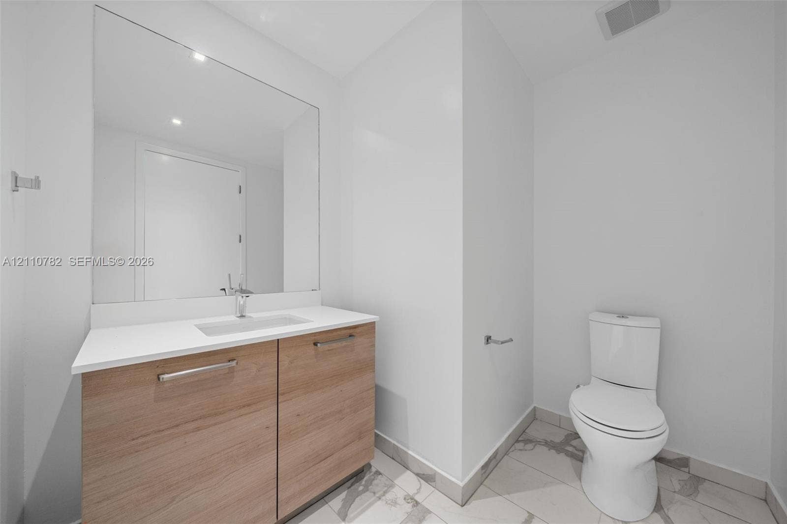 Vanity featuring a light wood-finish cabinet, white countertop with an integrated rectangular sink, large wall-mounted mirror, and tile flooring with a veined pattern