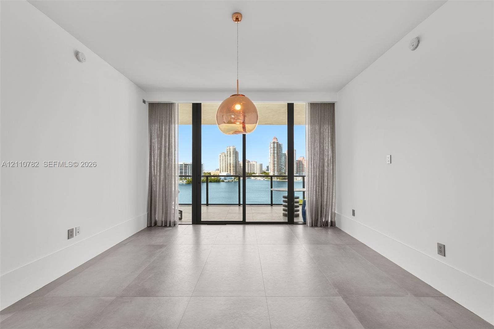 Large-format tile flooring, oversized sliding glass door, spherical glass pendant light, and balcony with water and city skyline backdrop