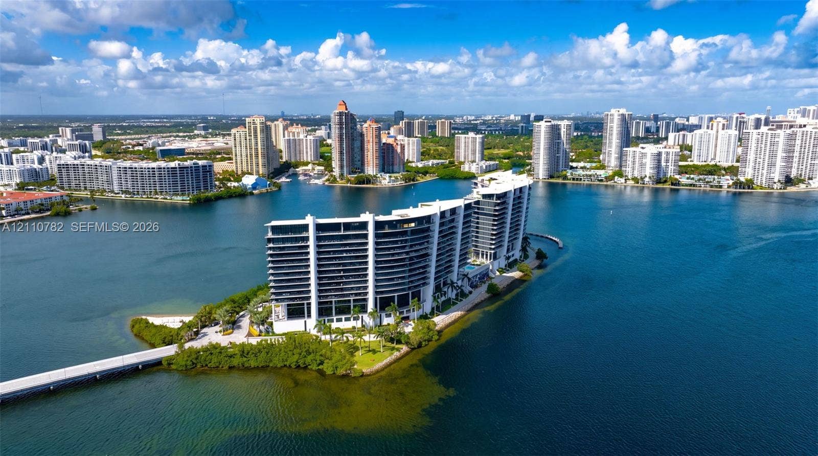 Waterfront high-rise building situated on a private peninsula, featuring wraparound balconies, an outdoor swimming pool, lush landscaping, and direct access via a private bridge