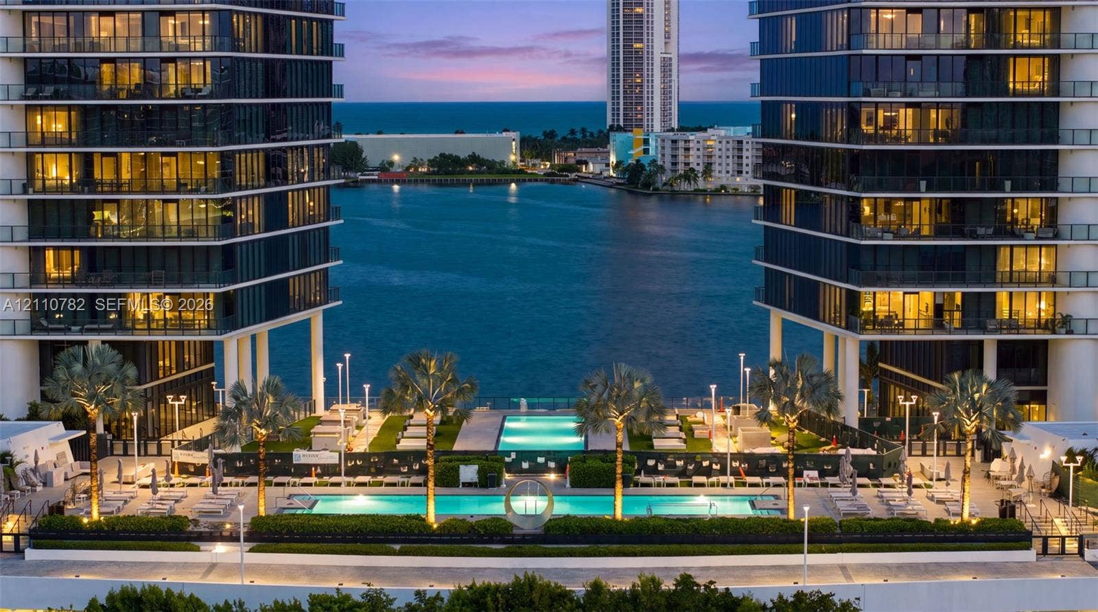 Waterfront high-rise complex featuring dual glass-facade towers, multiple swimming pools, professional landscaping, and paved outdoor terrace space