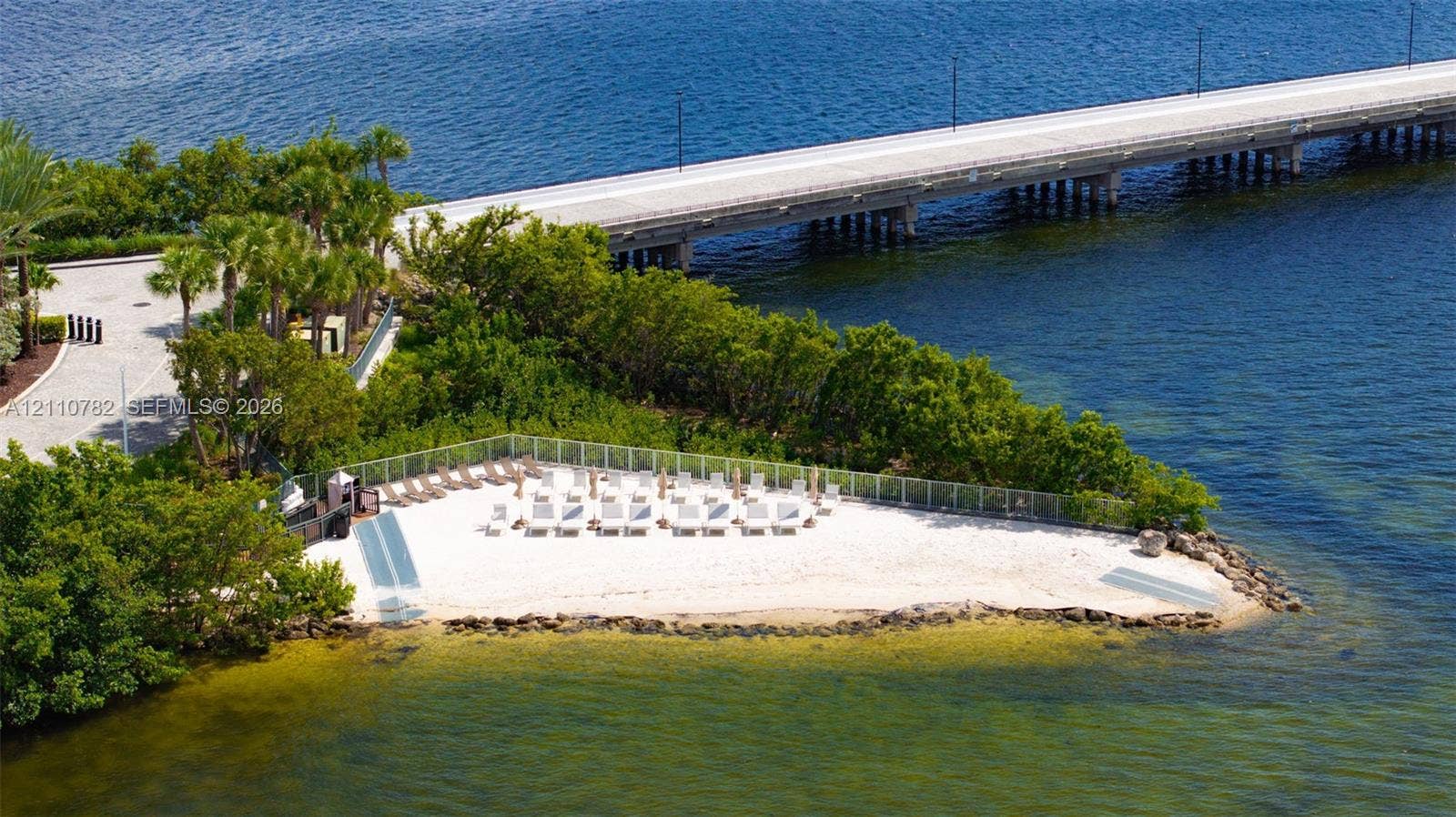 Private waterfront sandy beach featuring a perimeter fence, mature landscaping, and stone bulkhead