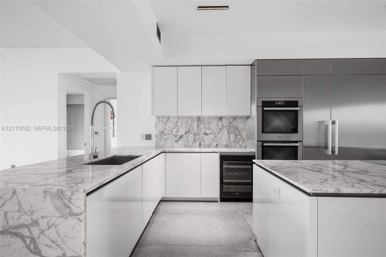 Kitchen featuring white flat-panel cabinetry, stone countertops with waterfall edge, full-height stone backsplash, stainless steel double wall ovens, and an under-counter beverage cooler
