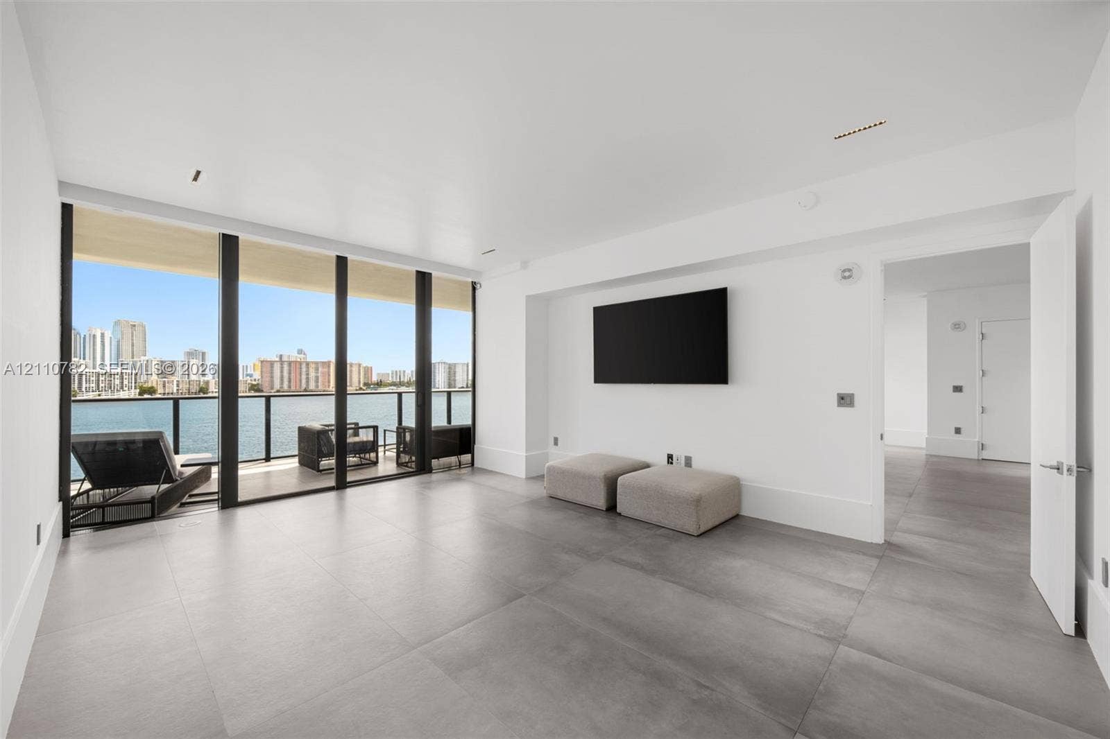 Large-format tile flooring, floor-to-ceiling glass paneling, and an attached balcony with metal railing overlooking the waterfront and cityscape