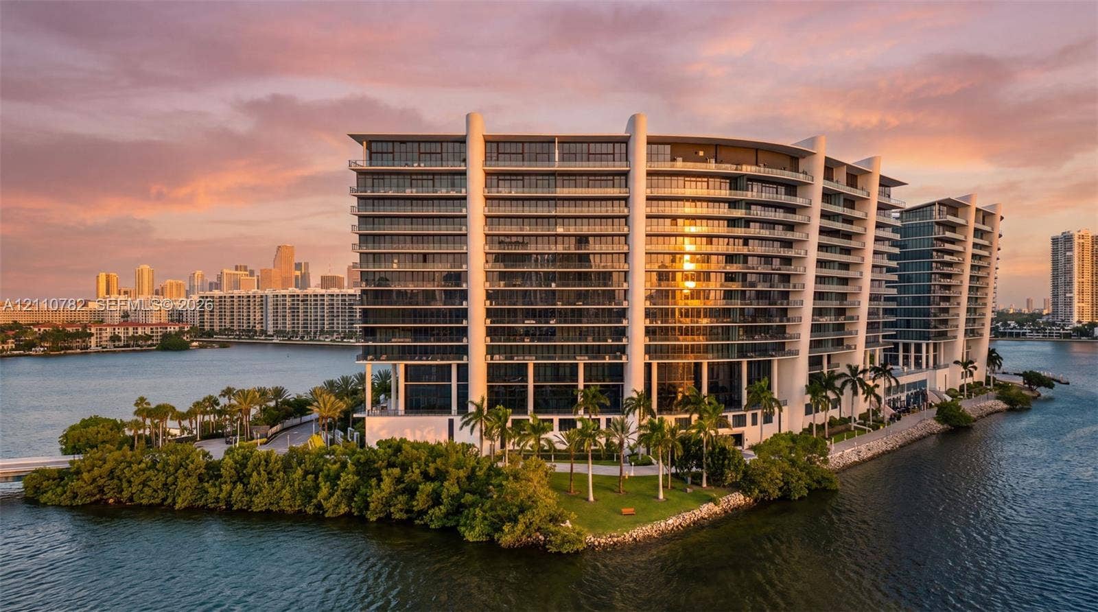 High-rise residential structure featuring tiered floor plans, expansive wrap-around balconies with glass railings, white structural pillars, and a waterfront location surrounded by mature palms