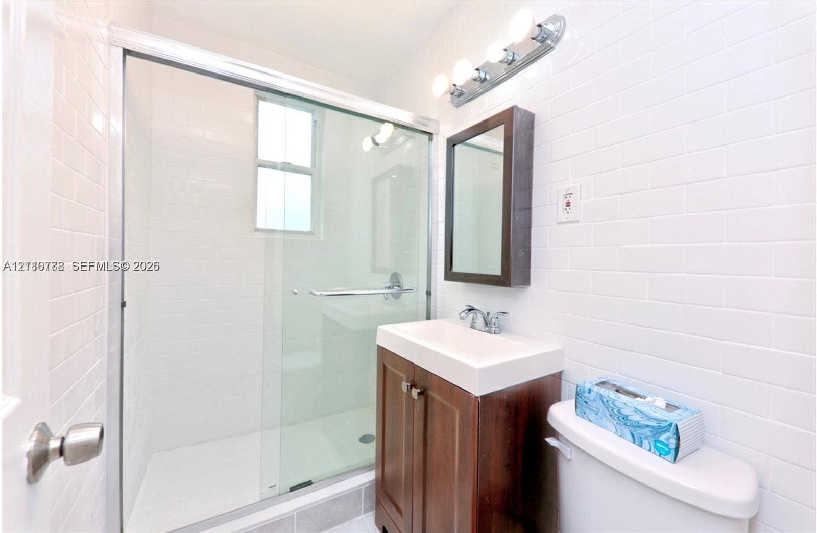 3710 Collins Avenue, #N-202, Miami Beach, FL 33140 Bathroom featuring a glass-enclosed shower with subway tile surround, wood-finish vanity, rectangular wall-mounted medicine cabinet, and four-bulb vanity lighting