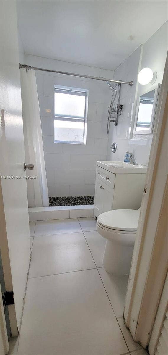 Bathroom featuring large-format floor tiling, white subway wall tile, a glass-enclosed shower with pebble-stone flooring, and a white vanity with integrated basin