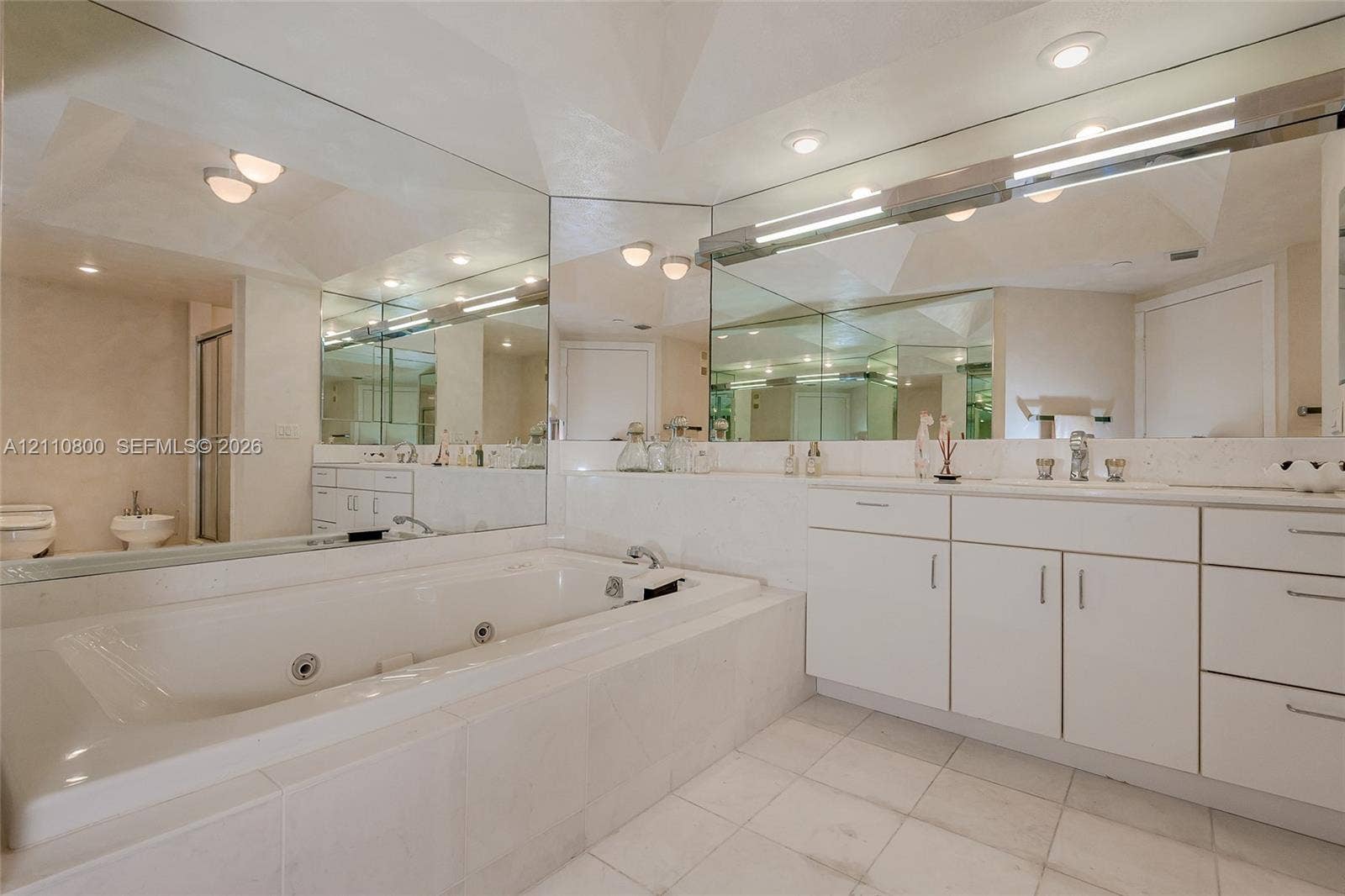 Bathroom featuring a large jetted soaking tub with a tile surround, white cabinetry, expansive wall-to-wall mirrors, recessed lighting, and tile flooring