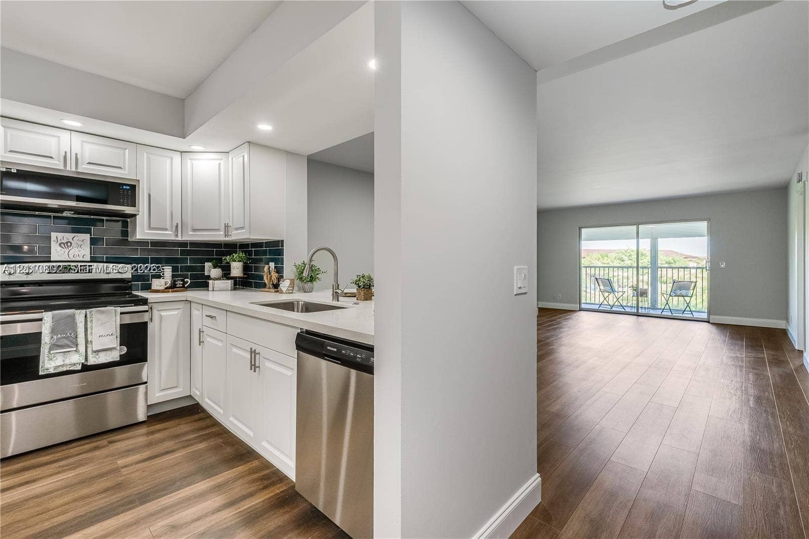 Kitchen featuring white shaker-style cabinetry, stainless steel appliances, dark tile backsplash, and light-toned countertops, complemented by wood-finish flooring extending into the common area with sliding glass doors