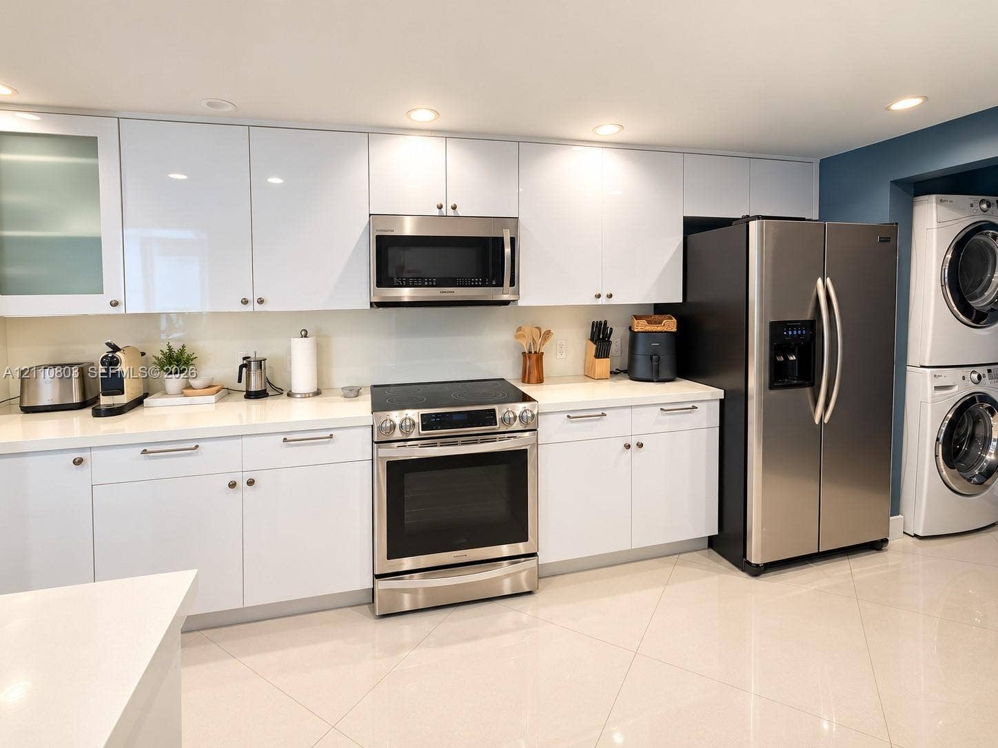 Contemporary kitchen featuring high-gloss white cabinetry, solid-surface countertops, stainless steel appliances, and large-format tile flooring, complete with a built-in laundry station