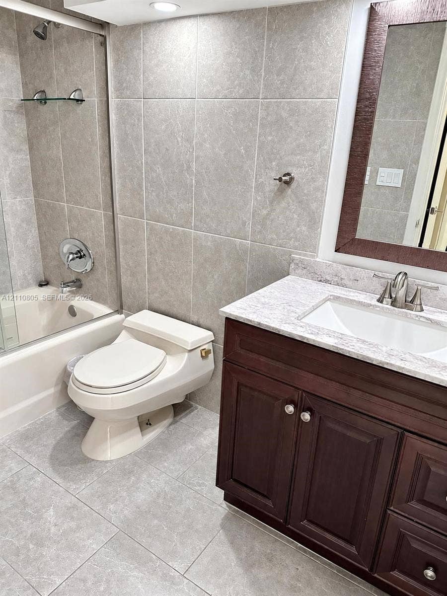 Bathroom featuring a dark wood-finish vanity with a stone-composite countertop, full-height tile wall surrounds, glass shower enclosure, and oversized ceramic floor tiling
