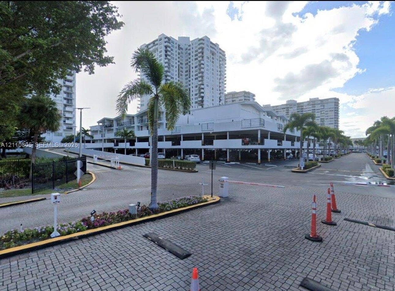 Covered parking structure featuring multiple levels, paved brick driveway, and manicured landscaping with palm trees