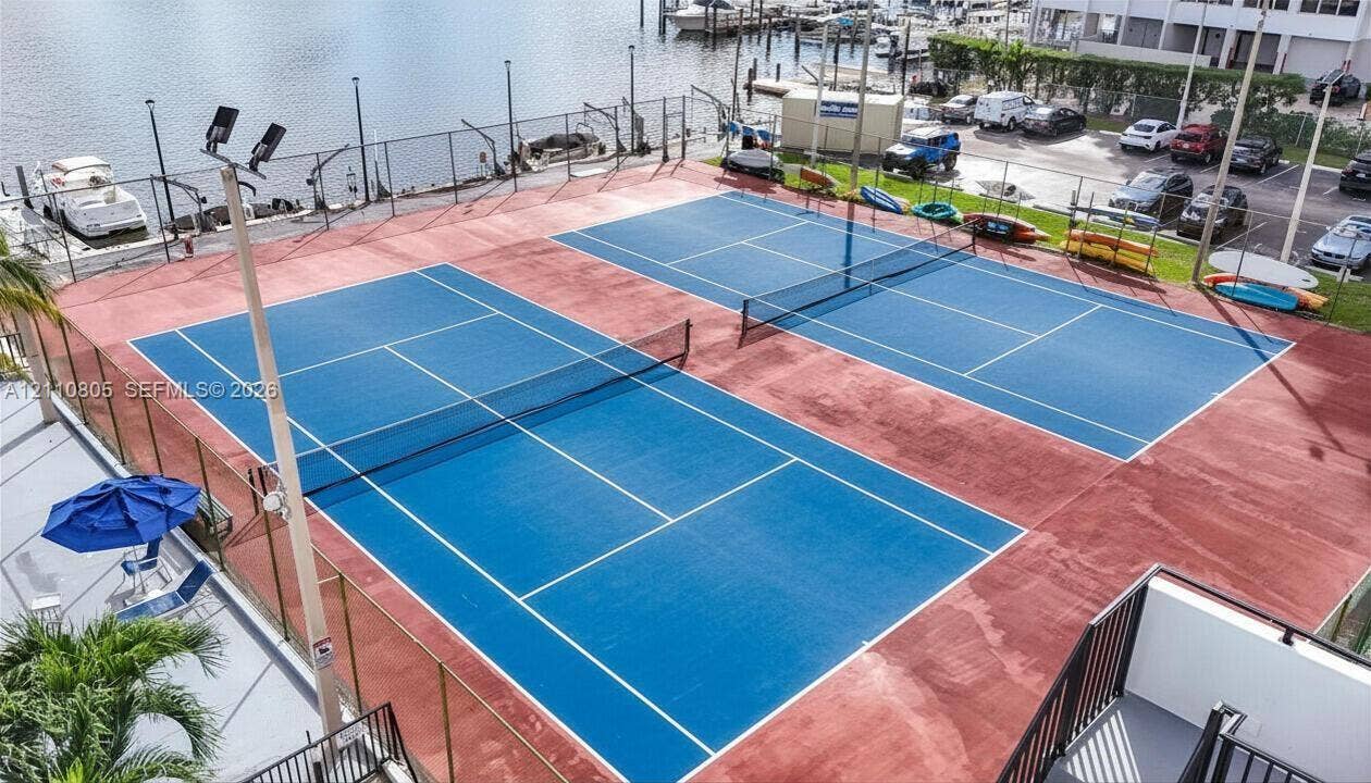 Dual tennis courts featuring high-contrast blue surfaces and perimeter chain-link fencing, situated waterfront with integrated lighting poles