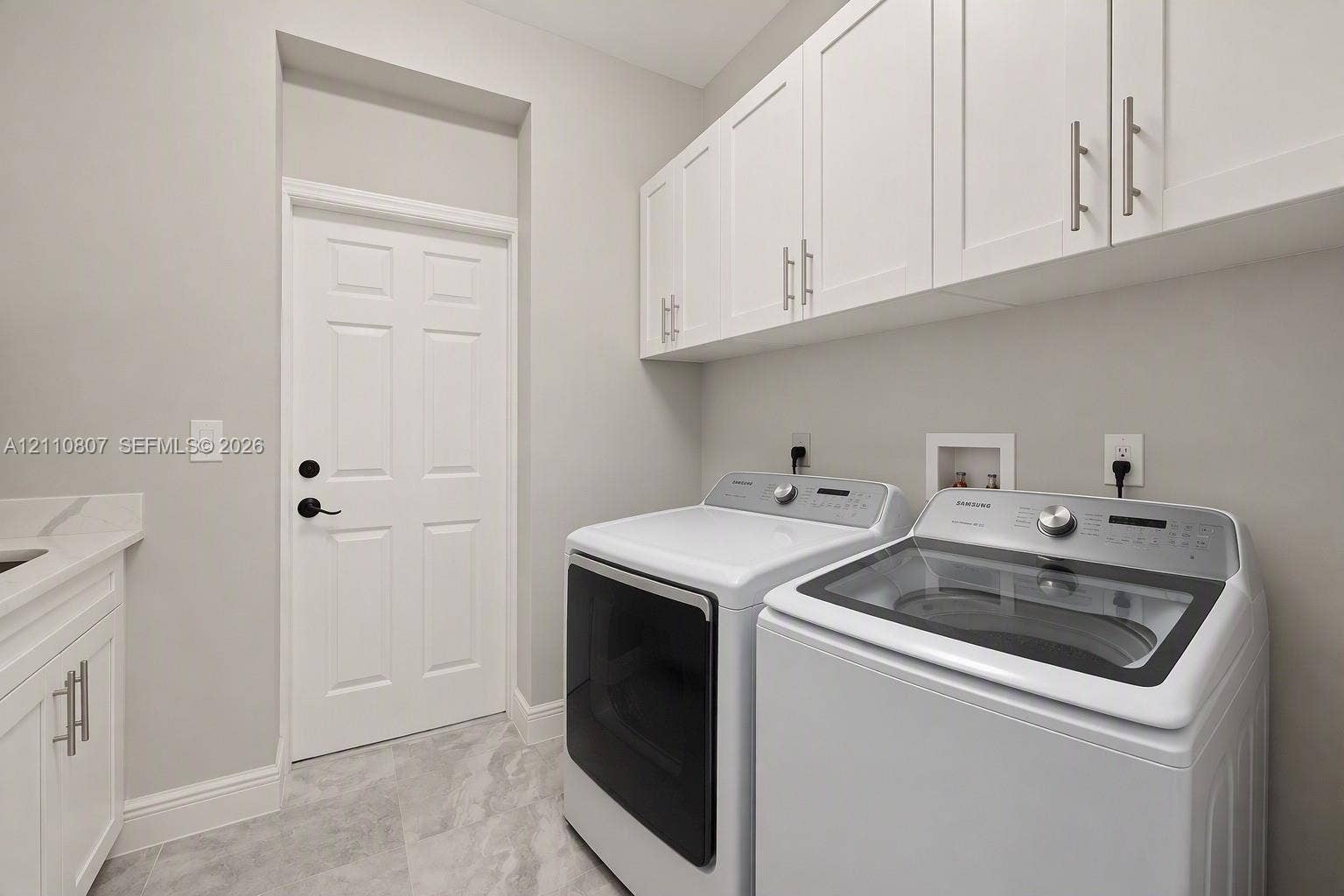 Laundry room featuring white Shaker-style upper cabinetry, stone-finish tile flooring, solid-surface countertop with integrated sink, and matte black door hardware