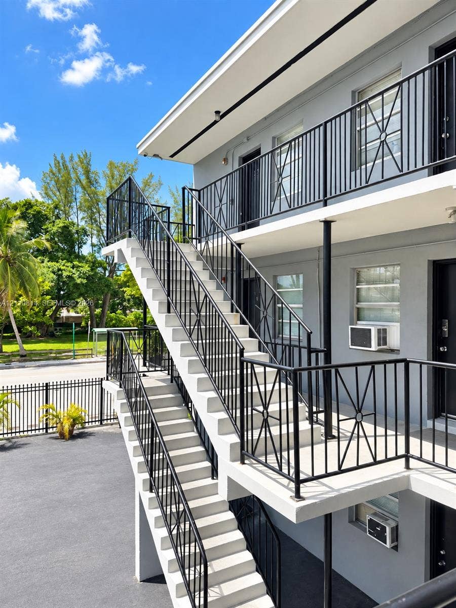 Multi-story exterior featuring a concrete staircase, black metal railings with geometric detailing, gray stucco finish, and paved ground-level parking