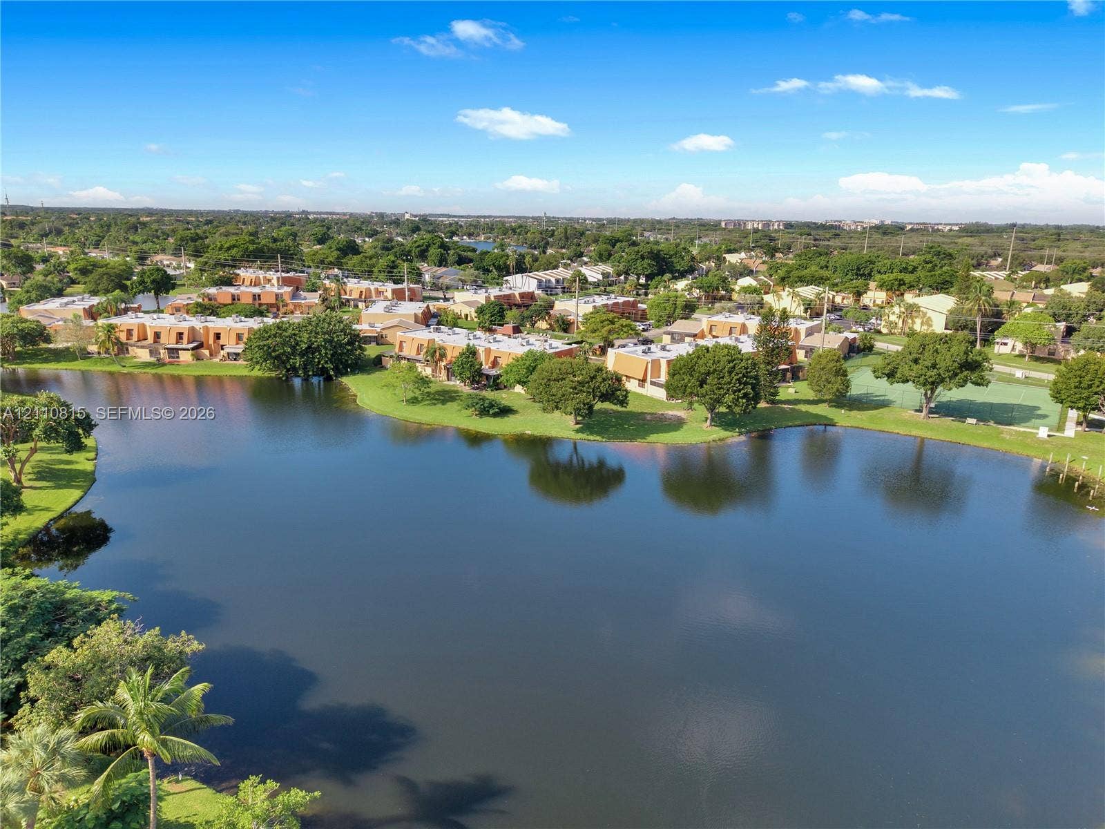 9100 Orchid Tree Lane, #137, Pembroke Pines, FL 33024 Aerial perspective of waterfront property featuring expansive lake frontage, mature trees, and an outdoor recreational court