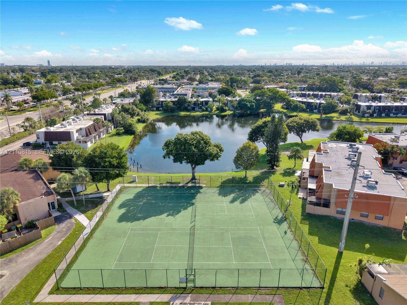 9100 Orchid Tree Lane, #137, Pembroke Pines, FL 33024 Aerial perspective of a fenced tennis court situated by a waterfront pond featuring mature landscaping and surrounding multi-unit residential structures