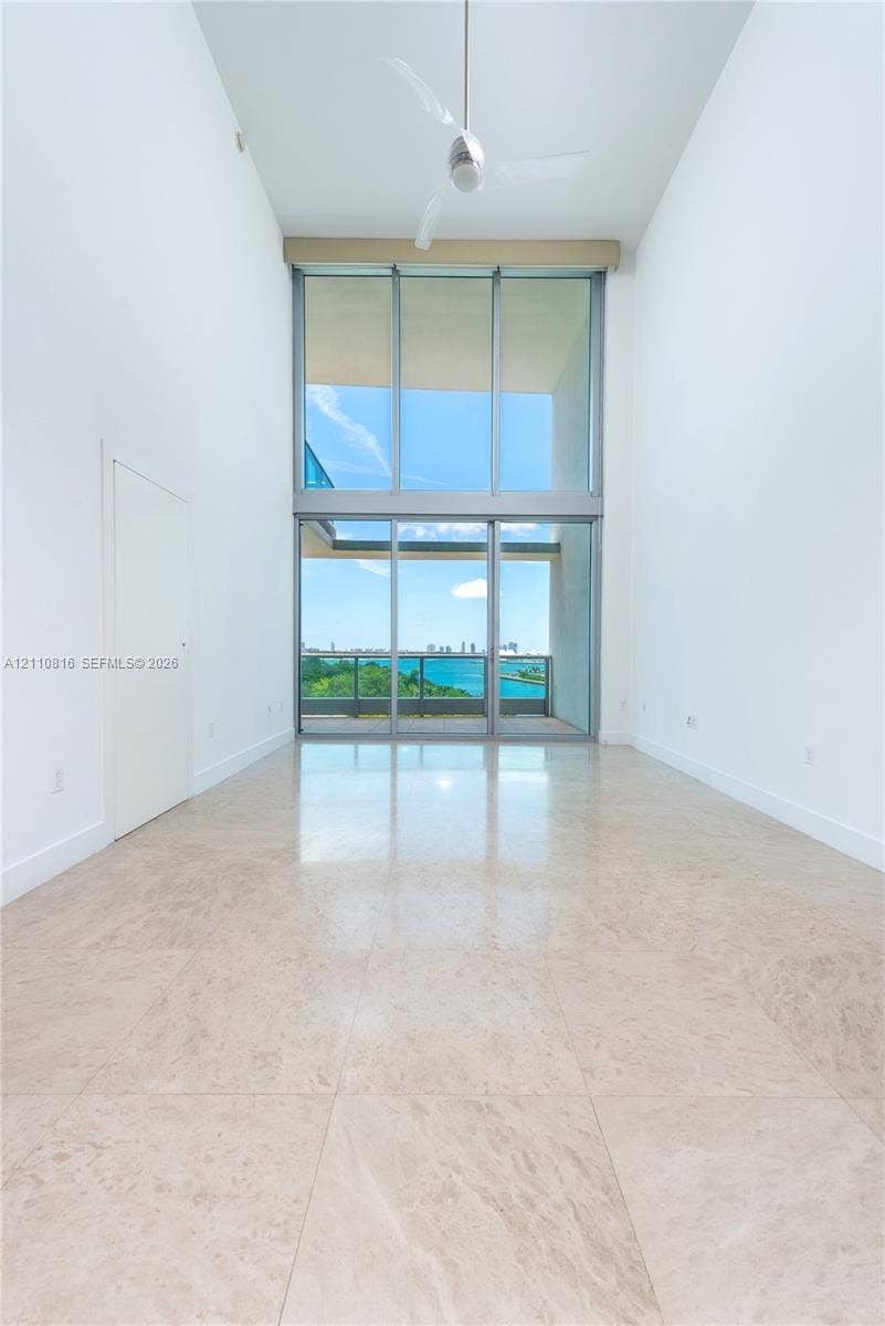 Double-height ceilings, floor-to-ceiling glass wall, polished stone flooring, and ceiling-mounted fan, overlooking water and city skyline