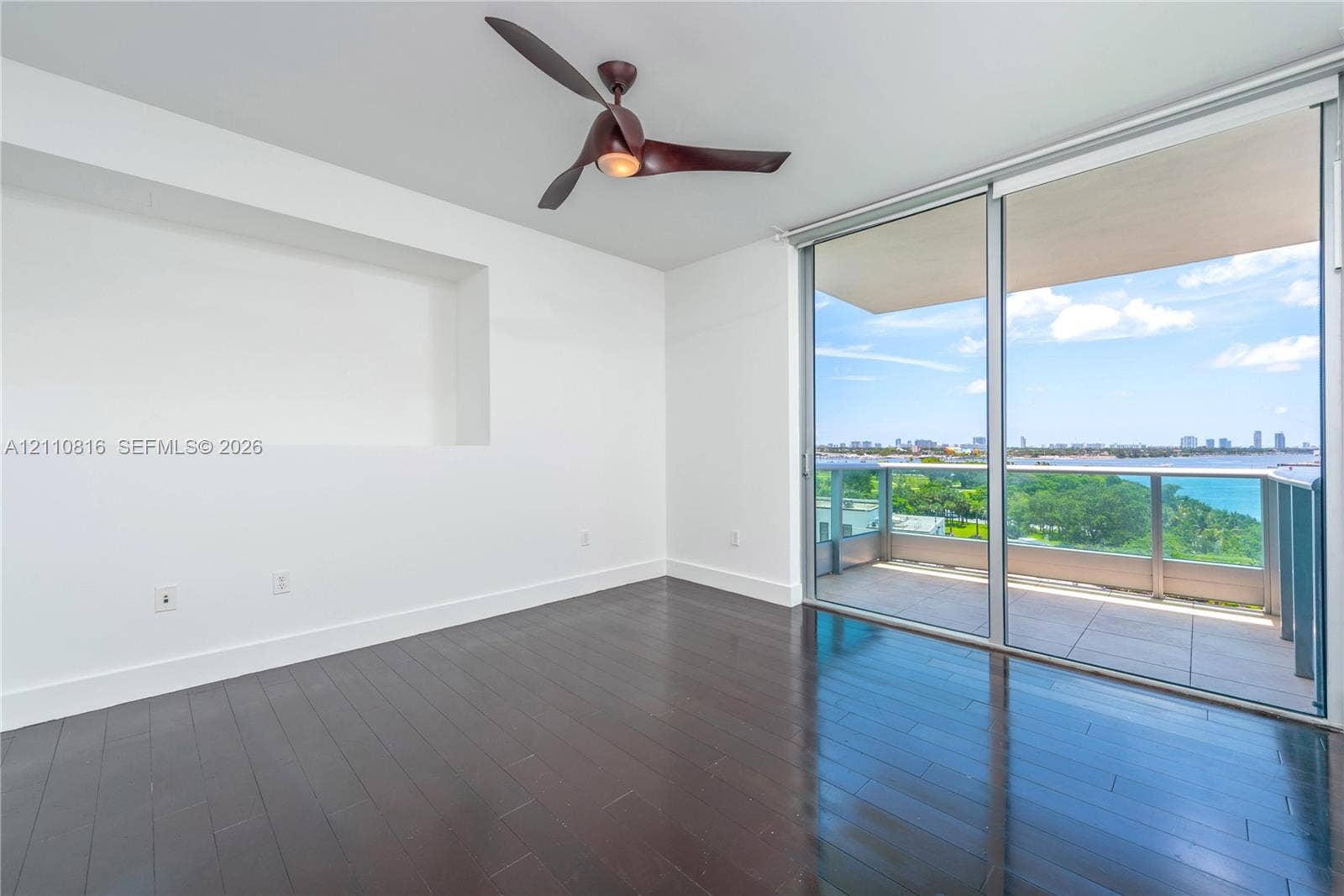 Dark wood-finish flooring, recessed wall niche, modern blade-style ceiling fan, and floor-to-ceiling sliding glass doors opening to a private balcony with glass railing