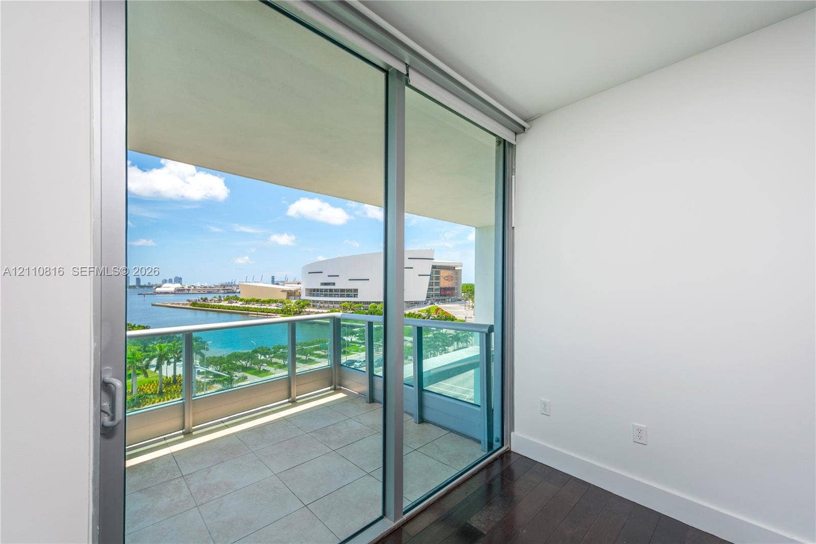 Floor-to-ceiling sliding glass door access to a covered balcony with a metal and glass railing, featuring tile flooring and hardwood-finish interior flooring